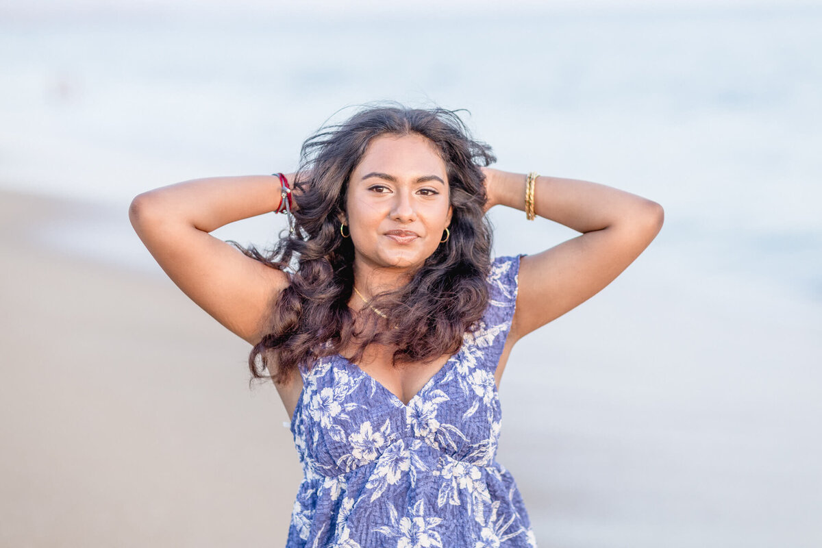 A person stands on a sandy beach with their hands behind their head. They wear a blue and white floral dress and have long, curly hair. The ocean is visible in the background.