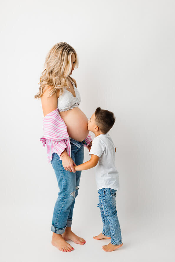 A boy kisses his moms bare, pregnant belly in a studio setting.