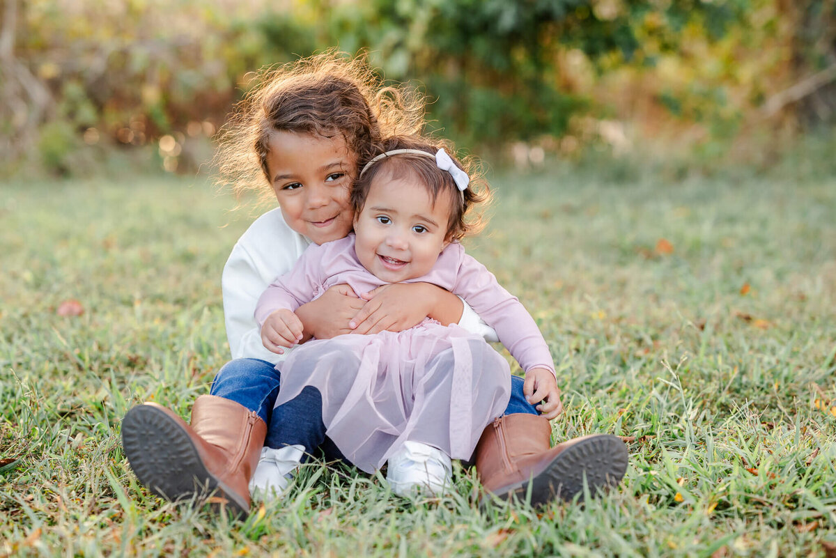 A young girl holds her toddler sister in her lap. They both smile for the camera while sitting in the grass .