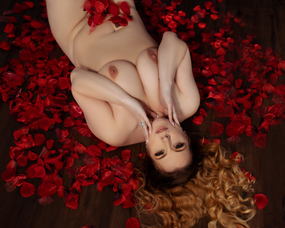nude woman with curled hair laying on black sheet set with red rose petals on her body and around her, boudoir
