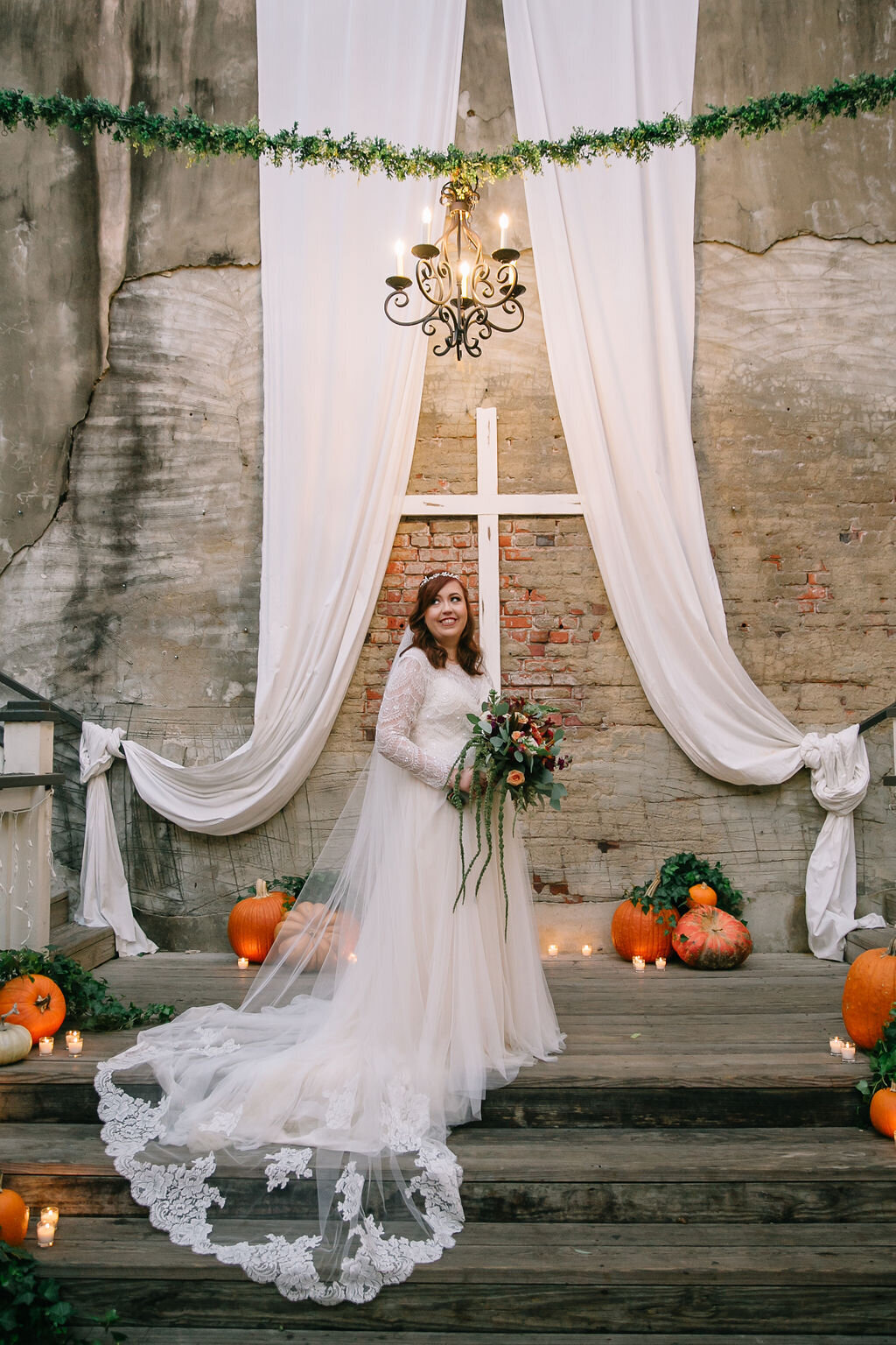 A smiling bride in a long-sleeved lace gown holds a bouquet, standing on wooden steps with pumpkins, candles, and greenery, in front of a cross, draped white fabric, and a brick wall.