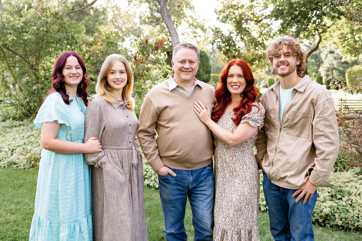 A smiling family of five posing in a lush green garden, dressed in coordinated neutral tones, with vibrant foliage in the background.