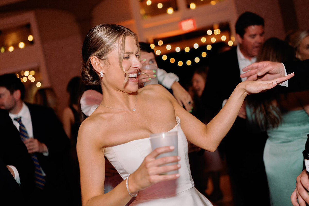 Bride laughing and dancing with a drink in hand during the lively wedding reception at Burritt on the Mountain, with guests and string lights in the background.