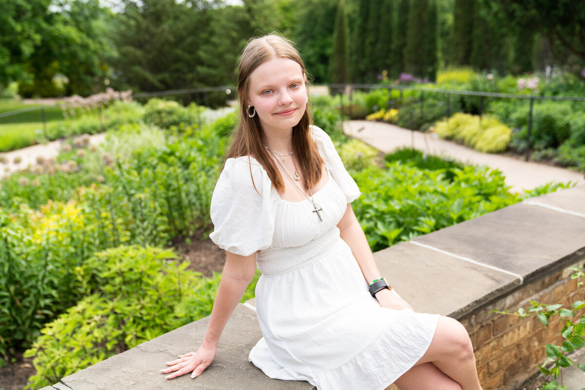 Senior girl wearing a white dress sits on a ledge during her senior pictures at the Minnesota Landscape Arboretum.