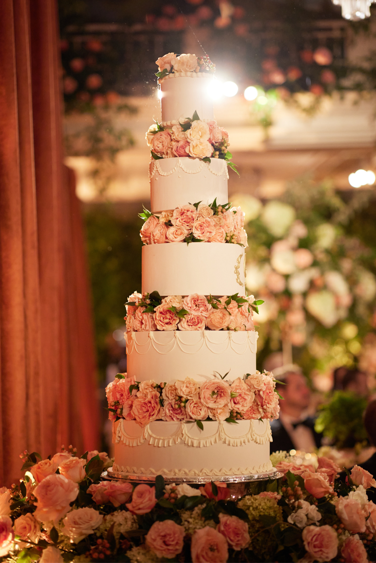 The photo was taken at the Willard Intercontinental, one of the top wedding venues and hotels in Washington D.C., by photographer Shawn Connell. Videographers Clayton Films captured the beautiful moment. The stunning image features a tall, multi-tiered wedding cake adorned with elegant white icing and cascading pink and white roses. The cake is beautifully showcased among a lush floral arrangement and set against a warmly lit, ornate backdrop, creating a truly picturesque scene.