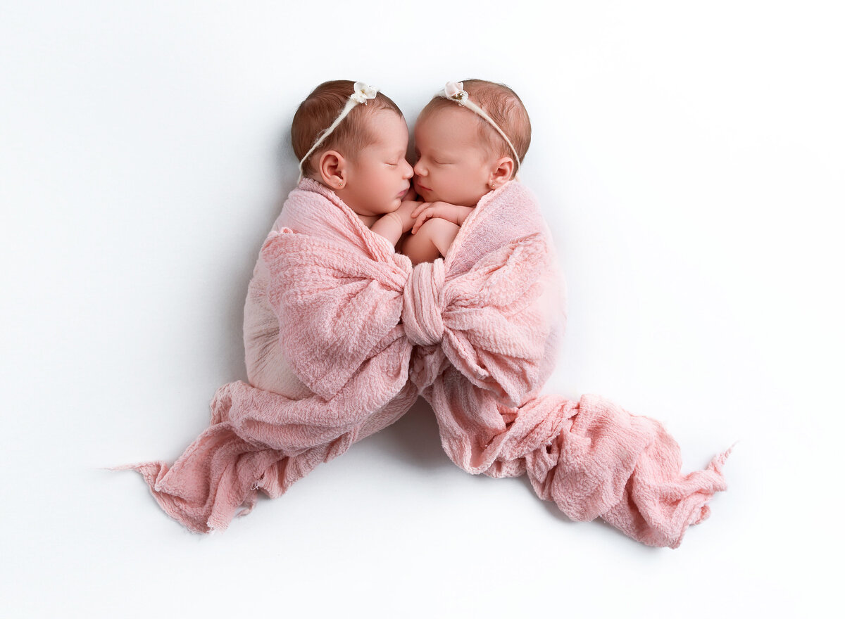 Twin baby girls are photographed in a Brooklyn, NY photoshoot. Baby girls are wrapped in a pink swaddle and tied to look like a large bow is trailing in fron tof them. Baby girls are holding hands and their heads are touching. Aerial image.