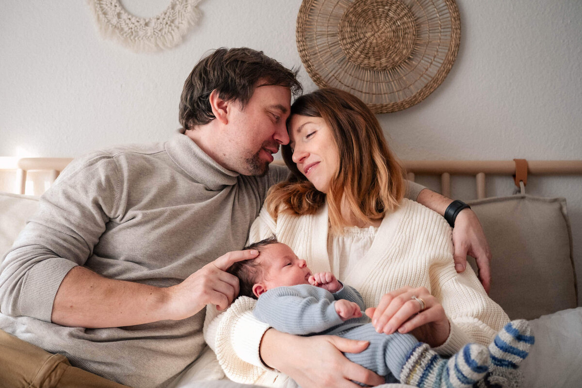 Parents sitting close with their foreheads touching as they hold their newborn baby in cozy knit clothing.