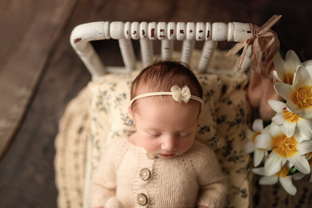 A beautifully styled newborn portrait taken at Meraki Photographic Creations in Paris, Ontario. Featuring a peacefully sleeping baby dressed in a hand-knit neutral sweater, resting on a vintage floral cushion with warm tones and rustic textures, surrounded by soft blooms.