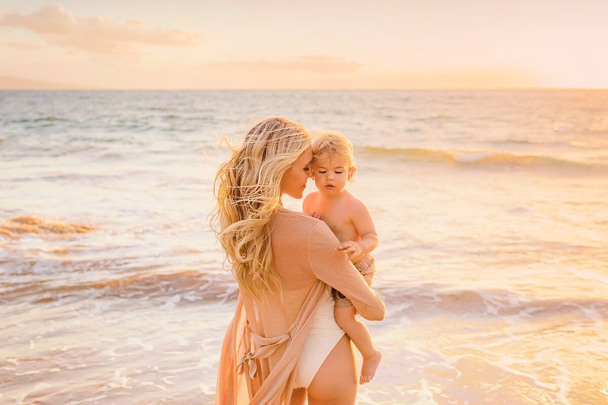 Mother holds her son at sunset on Maui next to the ocean as she walks toward the golden waves