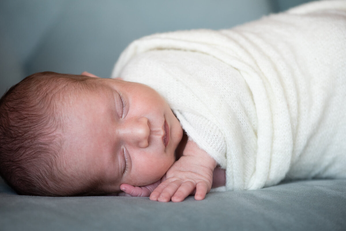 A newborn baby sleeps on its side on a blue chair while wrapped in a white swaddle with hands out as posed by a Denver Newborn Photographer