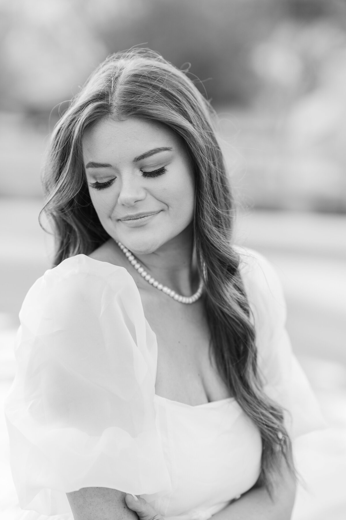 Black and White of Texas A&M senior girl looking over shoulder in a fluffy white dress and pearl necklace while sitting on edge of fountain in front of Kyle Field