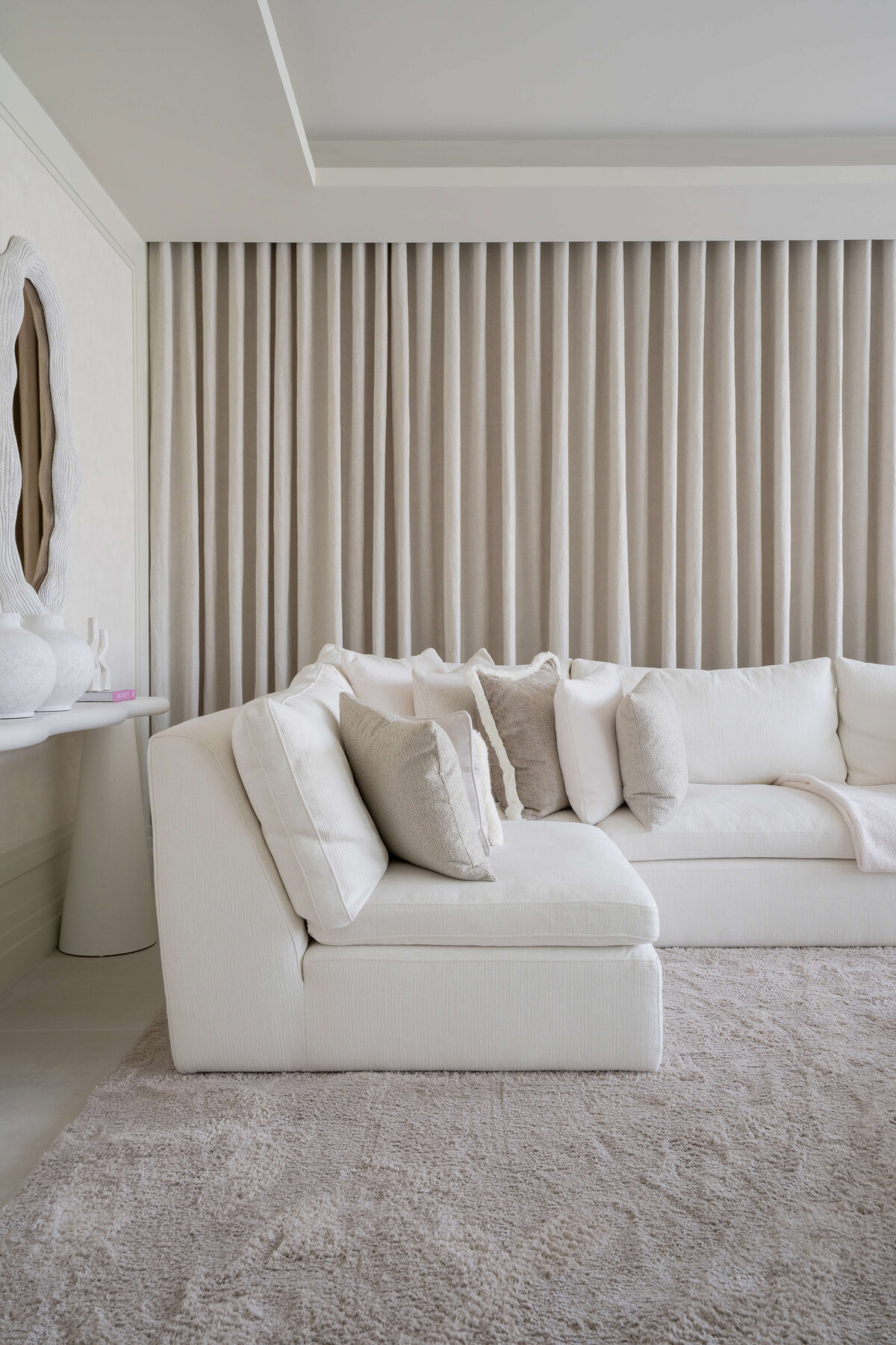 A spacious, modern living area. A large white couch with neutral throw pillows sits on a soft, textured cream rug. Behind the couch are floor-to-celiing cream drapes extending the length of the wall.