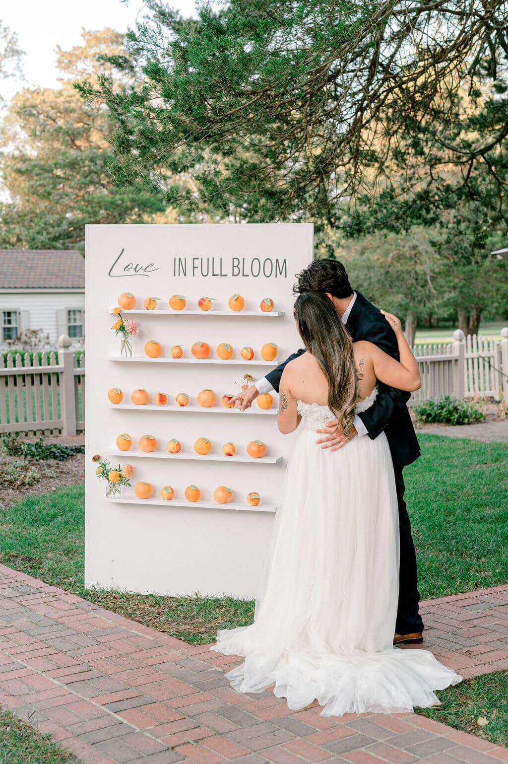 Bride and groom stand in front of a white escort display wall with oranges and floral accents that reads “Love in Full Bloom.”