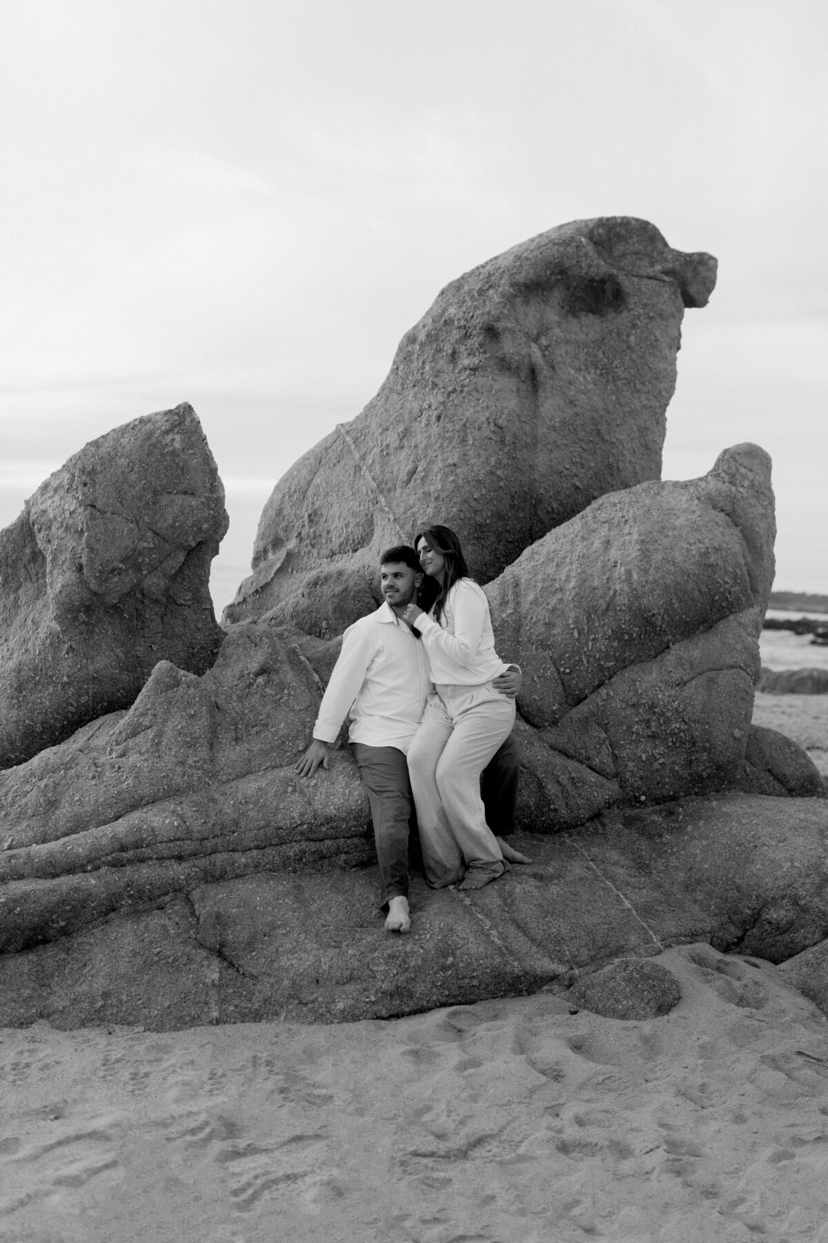 Black and white photo of man and woman sitting on big rock in white clothes on beach on coast in carmel california
