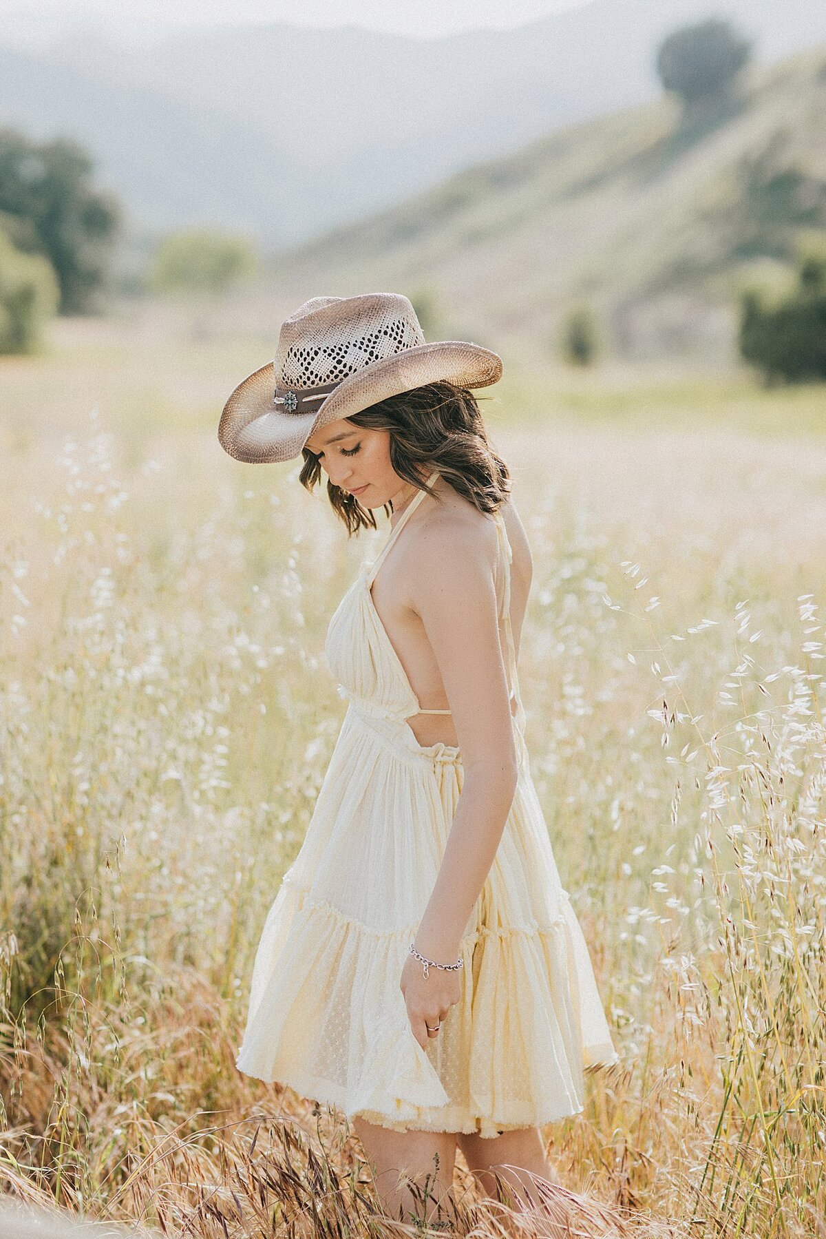 Boho-inspired senior girl in a straw cowboy hat and white crop top, standing in a wide open field with rolling hills behind her in Santa Clarita 