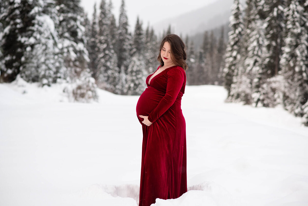 Brittany Danielle captures a Maternity image of a pregnant woman wearing a red velvet dress standing against a beautiufl snowy backdrop in Sun Peaks. She cradles her belly while she looks down at it.