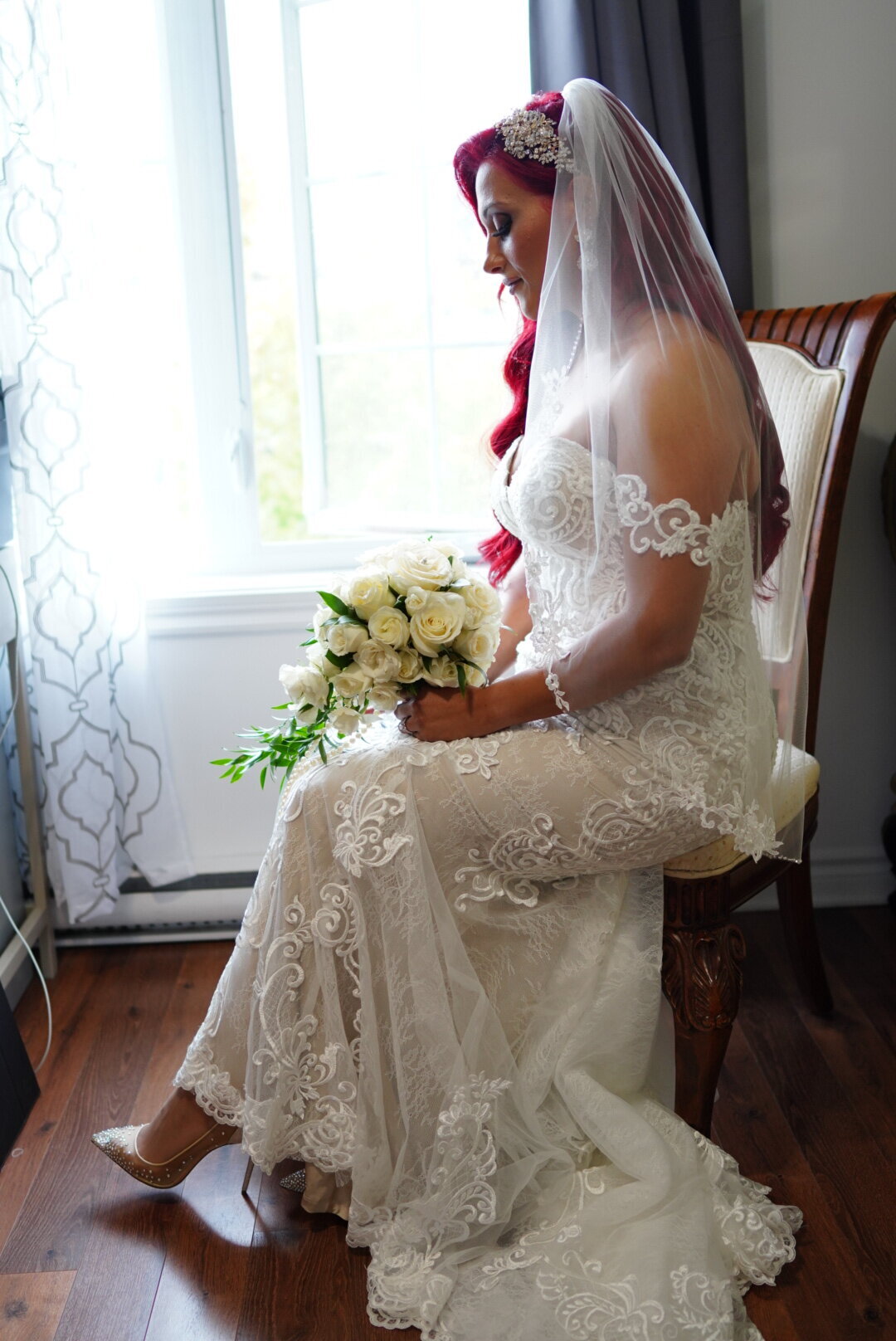 A bride sits elegantly by a window, bathed in soft natural light that enhances her radiant bridal gown. This image captures the bride’s serene beauty and the delicate interplay of light, creating a timeless and refined bridal portrait.