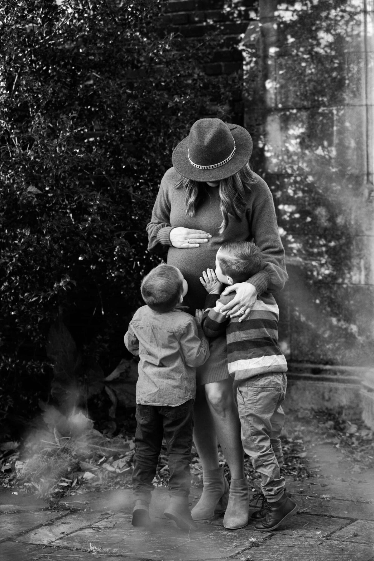 A pregnant woman wearing a hat stands while two young children affectionately touch her belly, captured beautifully by a maternity photographer from Pittsburgh.