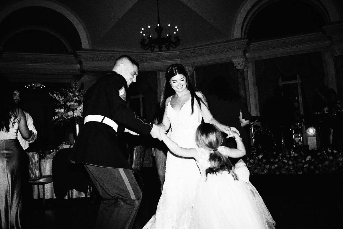 A man in a military uniform, a woman in a wedding dress, and a young girl dance together, smiling joyfully in a grand room—an unforgettable moment beautifully captured by an NJ wedding photographer.