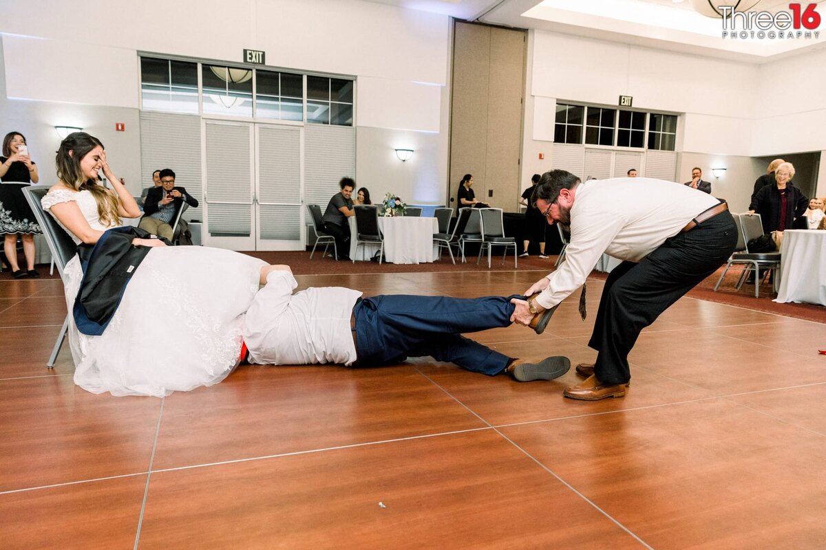 Father of the Bride pulls the Groom by the feet on the ground as the Groom is under his Bride's dress removing the garter belt as the Bride puts her hand on her forehead