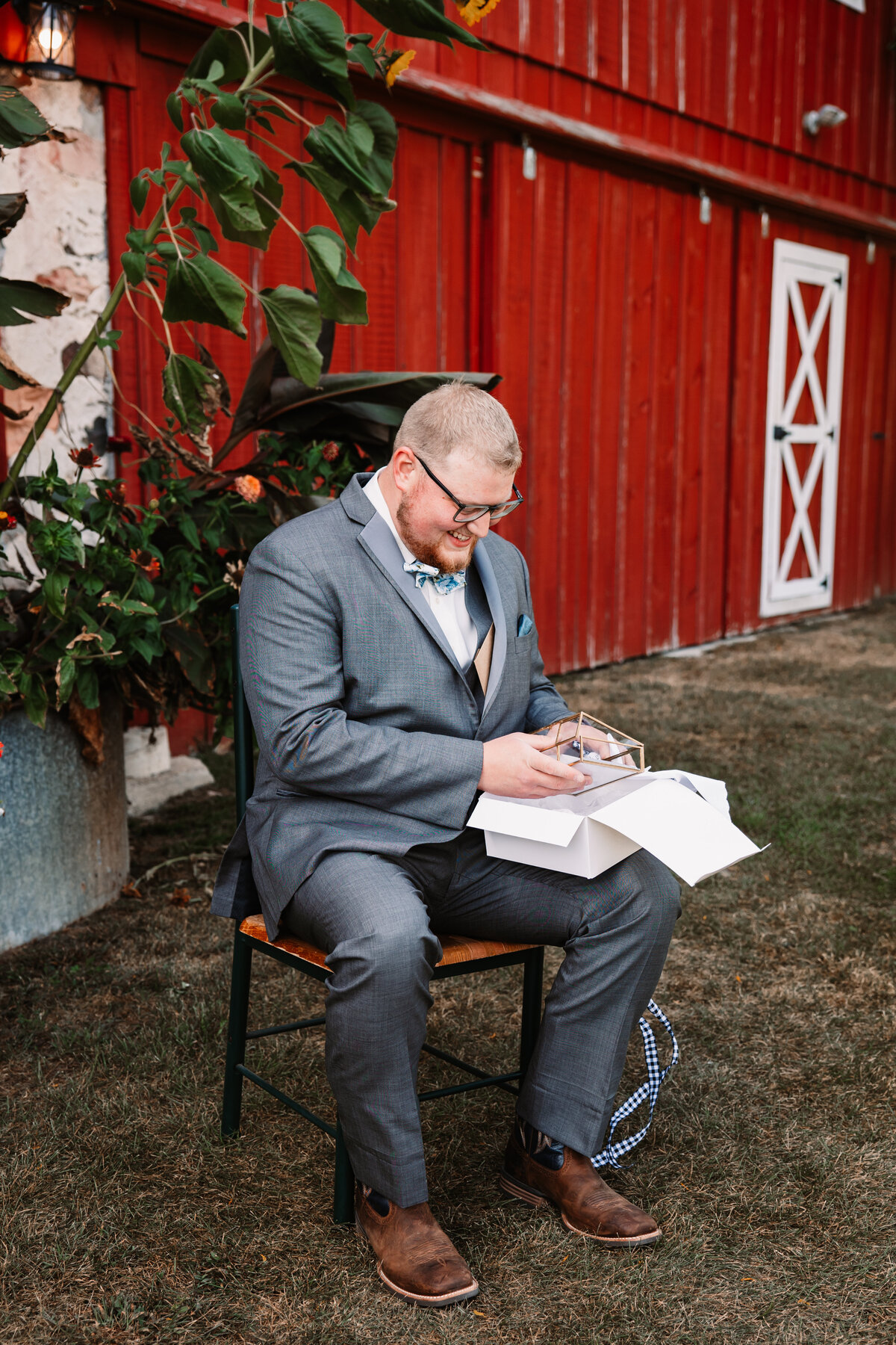 A groom in a grey suit sits in a chair opening a present in front of a red barn