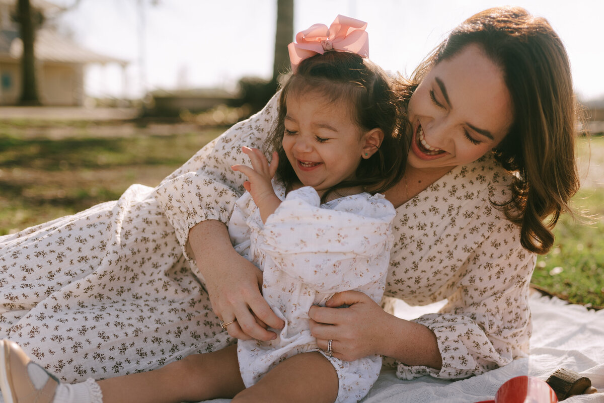 a mother tickling her daughter while sitting outside on a picnic blanket.