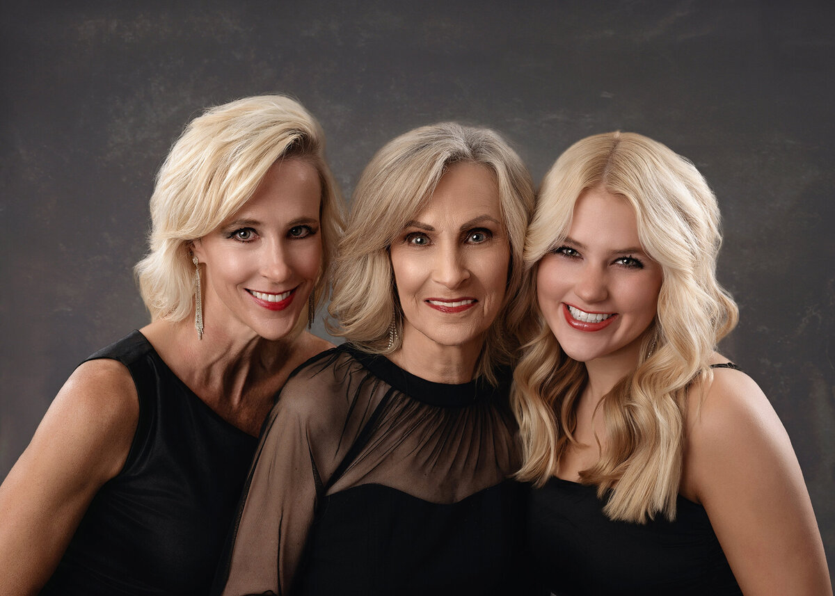 Three women from different generations with light hair are smiling and posing closely together against a dark, textured background.