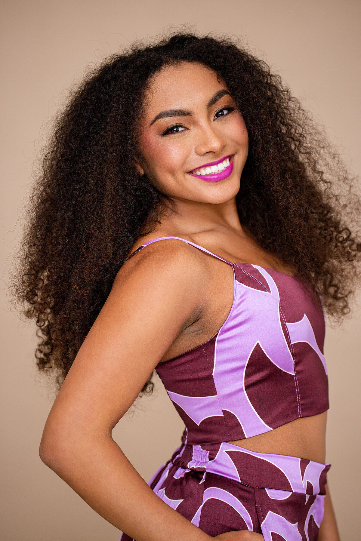 Confident teen girl with voluminous curls, smiling brightly in a purple and burgundy geometric two-piece set against a warm beige background.