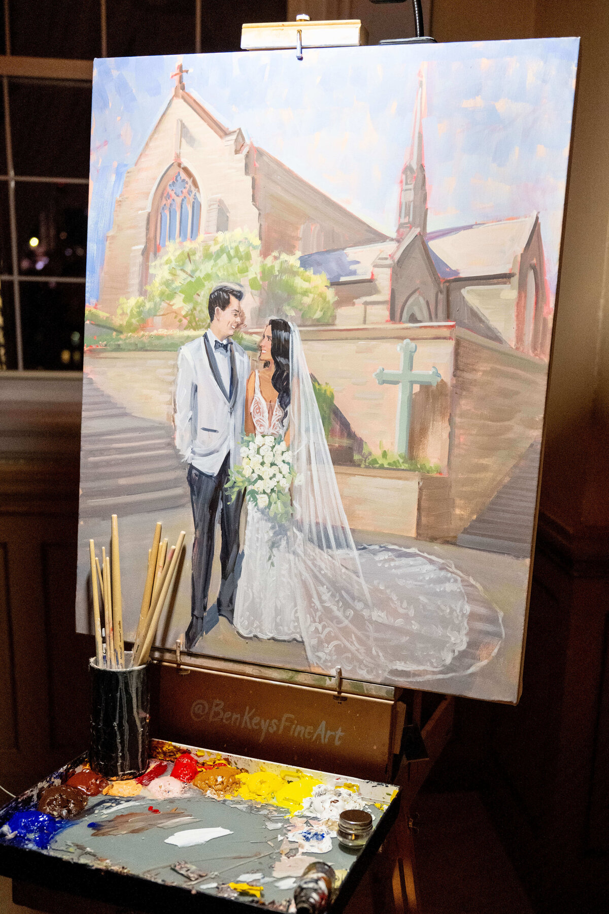 A painting on an easel depicts a bride and groom standing outside a church. The bride wears a white gown with a long veil, and the groom is in a suit. The scene is detailed with brushes and paints visible in the foreground.