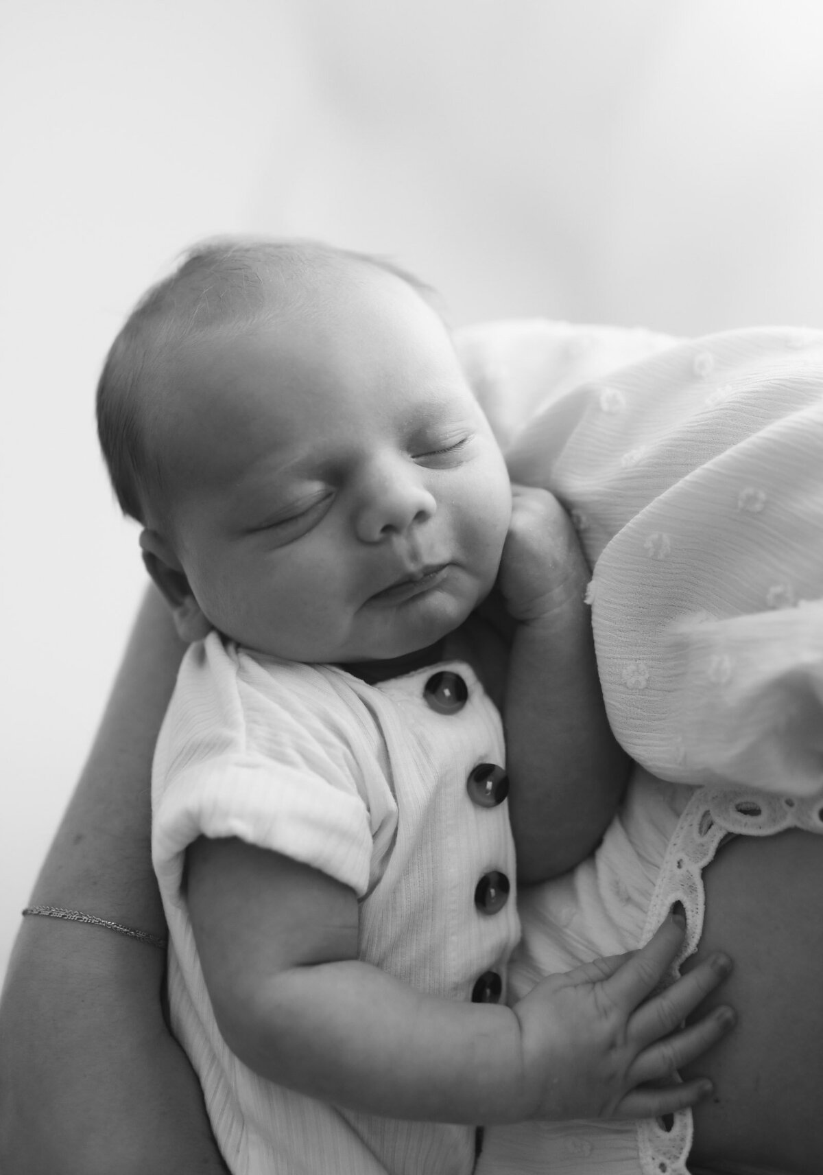 Black and white newborn photo of a baby sleeping and wearing a soft, white button-down onesie. The baby is in the arms of its mothers.