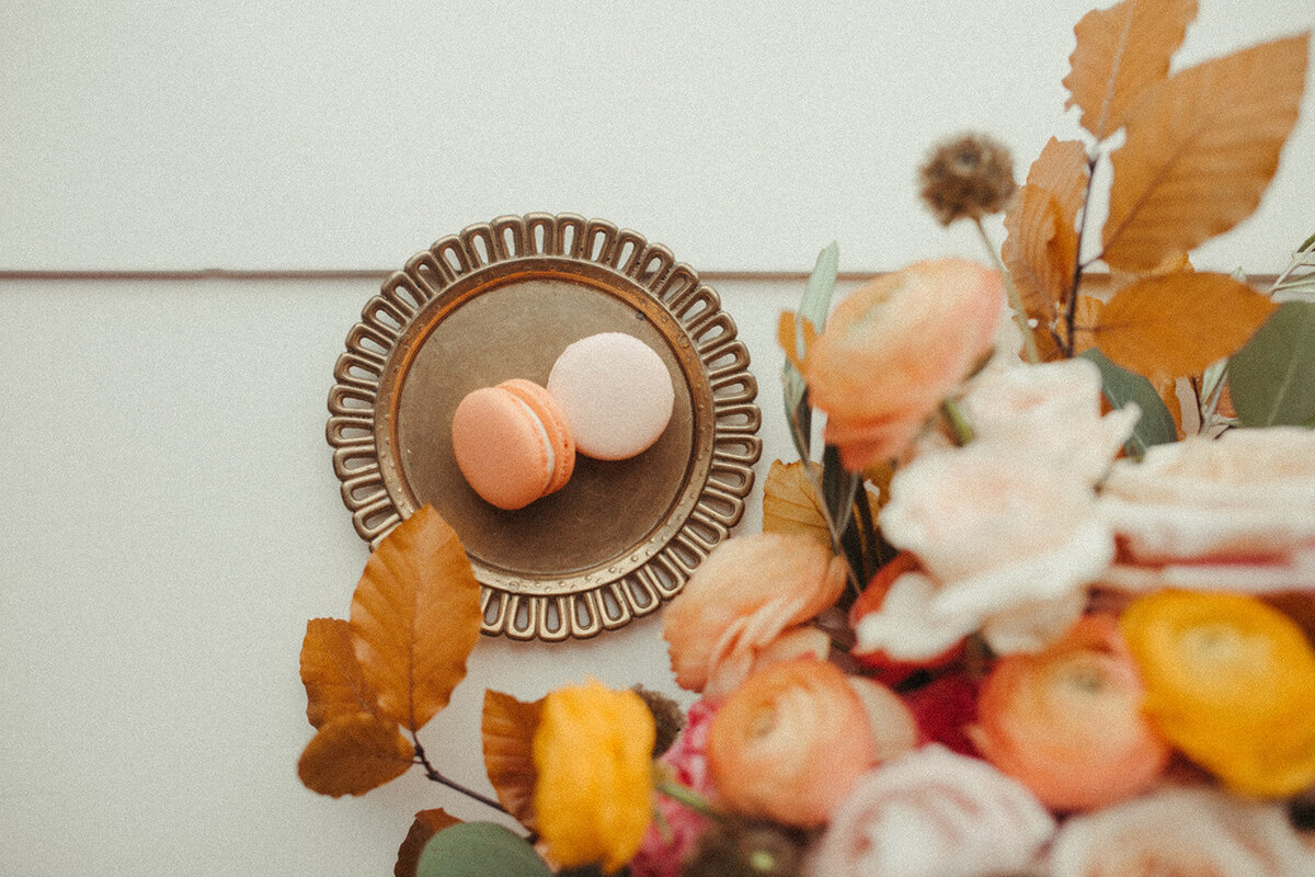 Two peach-colored macaroons on a bronzed dessert plate set next to an arrangement of flowers.