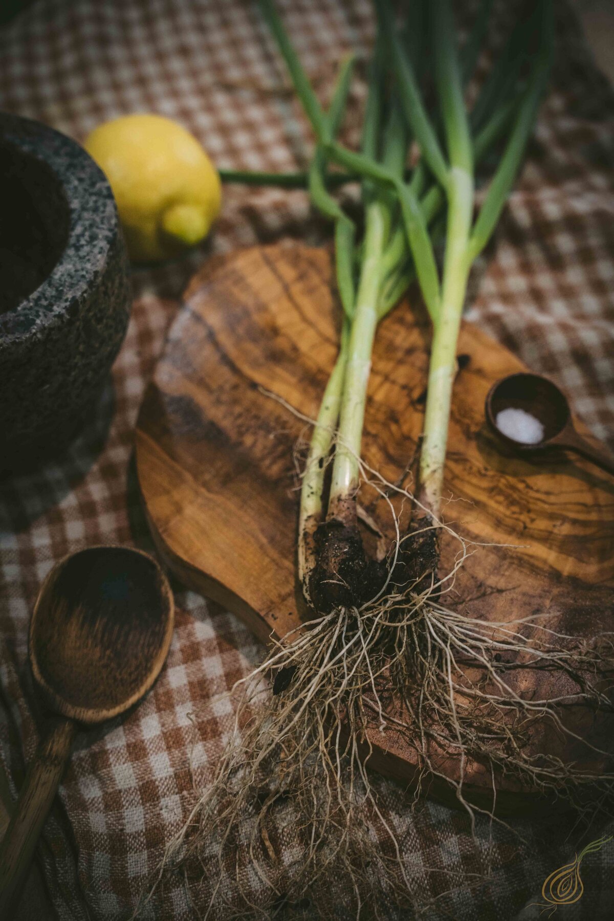 A wooden cutting board displays fresh green onions and a lemon alongside a mortar and pestle, highlighting ingredients for cooking.