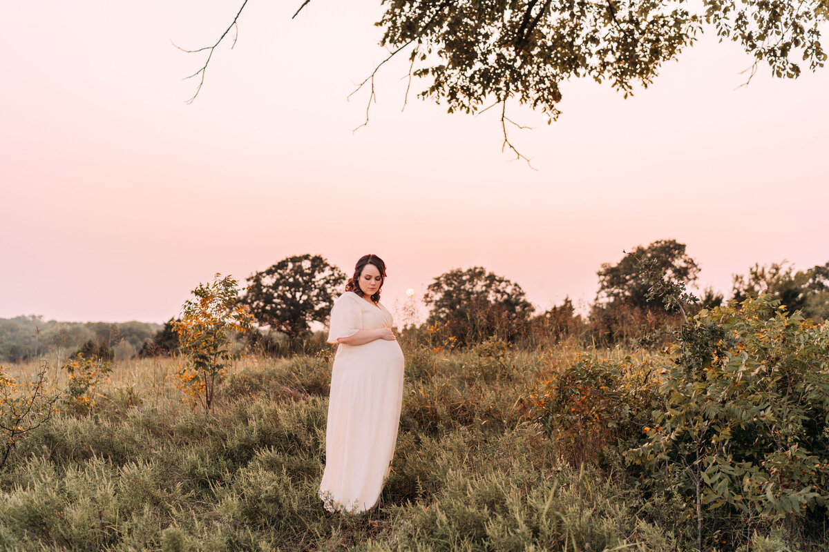 pregnant mother stands in white dress in fall field