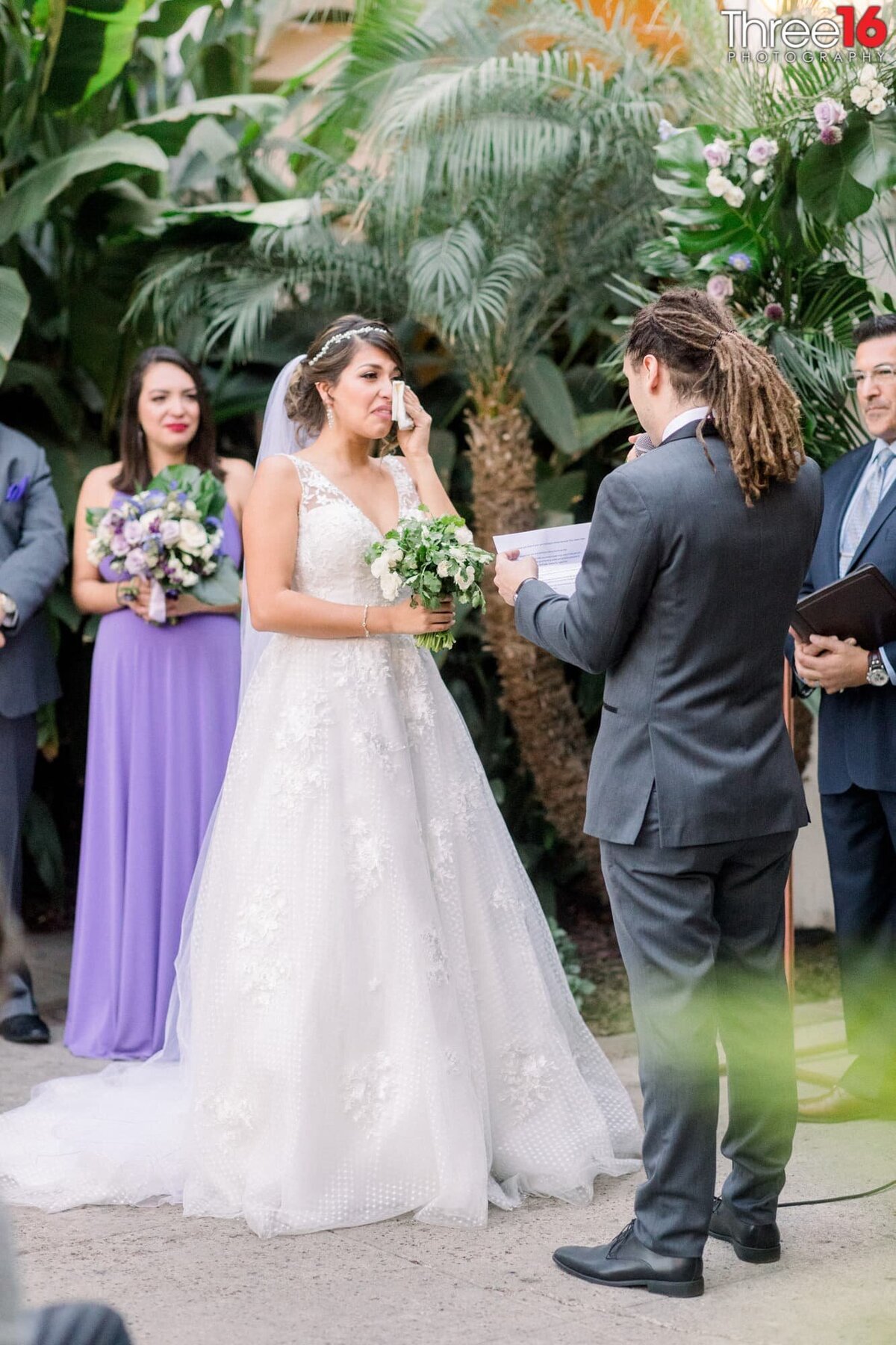 Bride wipes a tear from her cheek as her Groom reads his vows to her at the altar