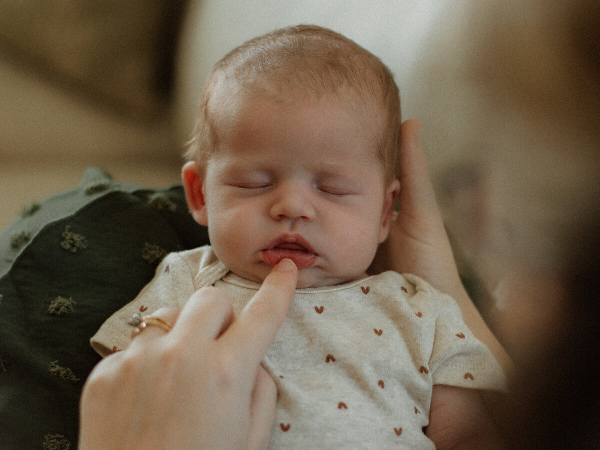 Newborn baby resting peacefully on a parent’s lap while a finger gently touches her lips.
