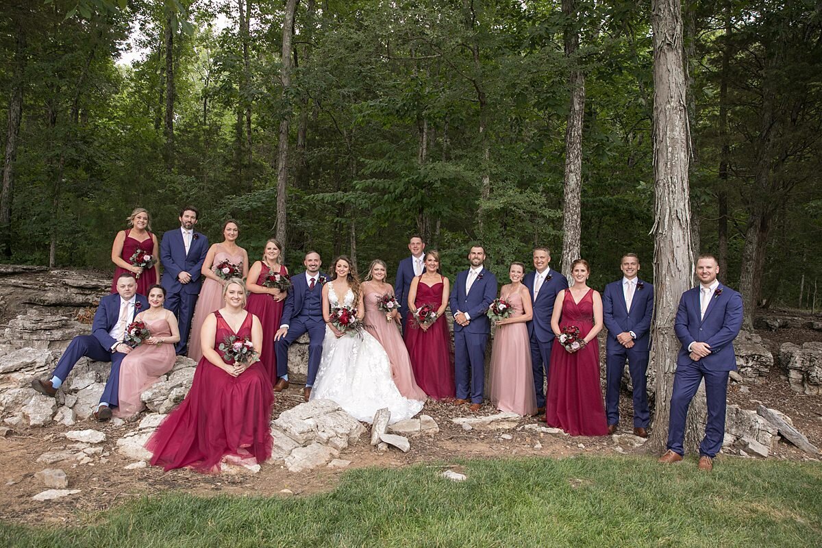 The bride, wearing a long sleeveless lace bridal gown with a plunging neckline, sits on a limestone rock outcropping in the woods with the groom and groomsmen dressed in navy suits and burgundy ties. The bridesmaids are dressed in blush bridesmaid dresses and burgundy bridesmaid dresses. They are holding bouquets of blush roses, burgundy roses, red peonies and burgundy ranunculus accented with greenery and wrapped in ivory satin ribbon.