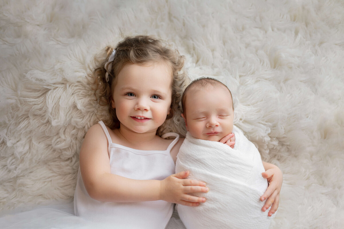 Newborn sibling photo with  a newborn baby and big sister holding him  on a white rug