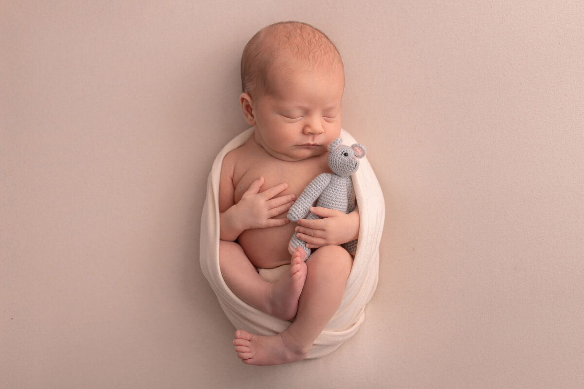 nyfödd bebis poserad på rygg på en beige bakgrund med beige wrap runt sig med en stickad mus i famnen