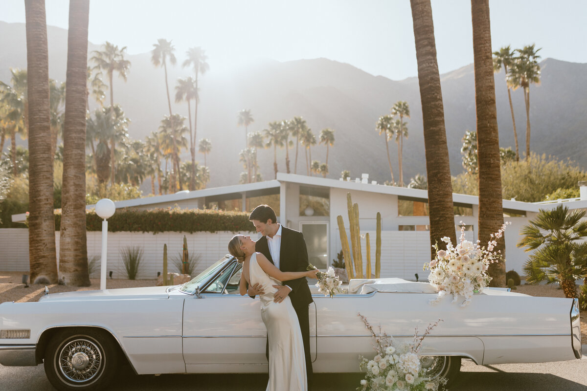 A newlywed couple embraces in front of a vintage white car adorned with flowers, surrounded by palm trees and mountains at sunset.
