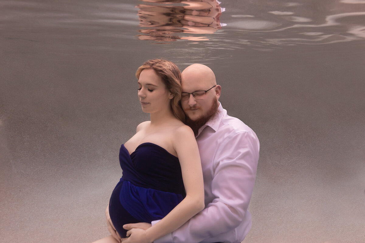 A tranquil underwater maternity photo captured by a Dallas underwater maternity photographer. A couple embraces tenderly beneath the surface, the man seated behind the woman with his arms wrapped lovingly around her, eyes closed in a moment of peaceful connection. The woman wears a flowing deep blue gown that gently curves around her belly, highlighting the grace and serenity of the scene. The soft, neutral background creates a dreamy, ethereal atmosphere, enhancing the feeling of intimacy and calm.