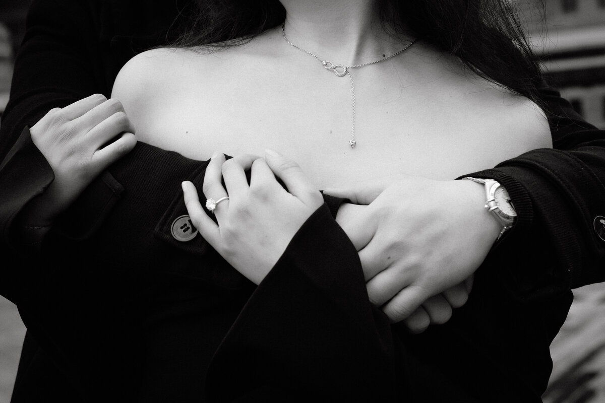 Black and white close-up of couple’s hands embracing, showing engagement ring and necklace during romantic NYC engagement session