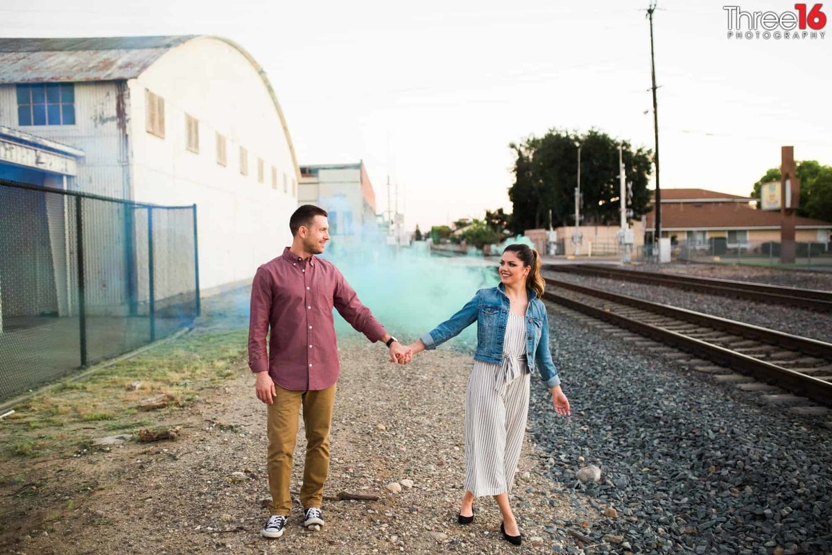 Engaged couple hold hands as the walk along the train tracks