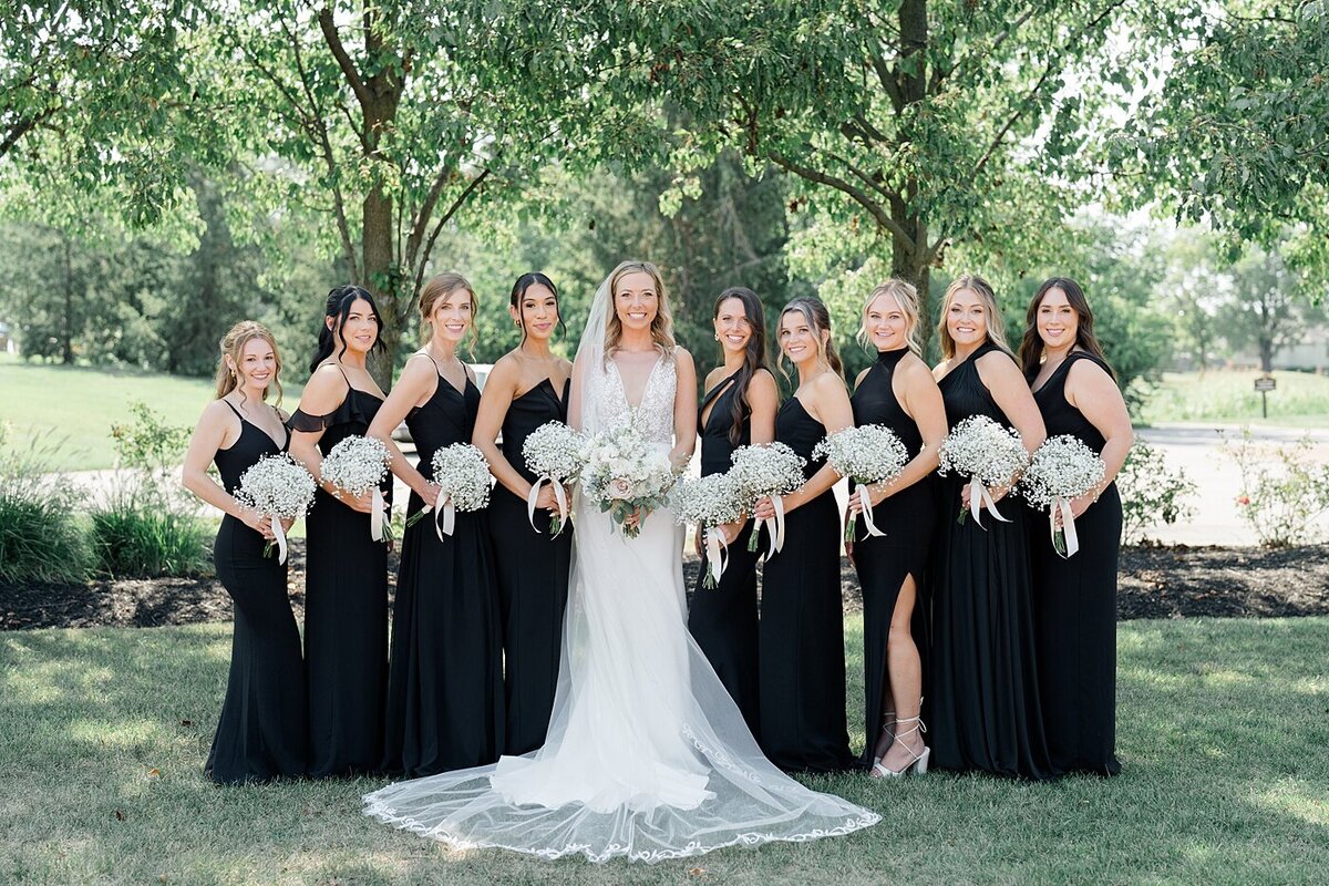 bride standing with her bridesmaids in a garden at the Pinnacle Golf Club with her bridesmaids in black dresses and holding floral bouquets