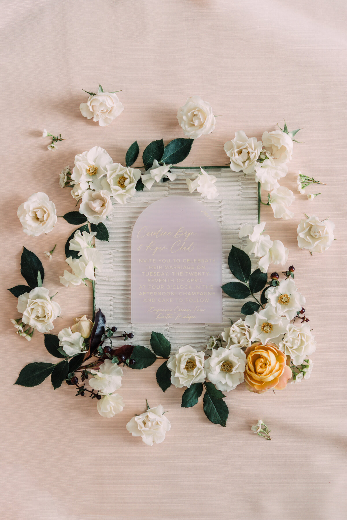 Styled wedding flatlay of frosted pink acrylic invitation on textured white fabric with white majolica spray roses and greenery