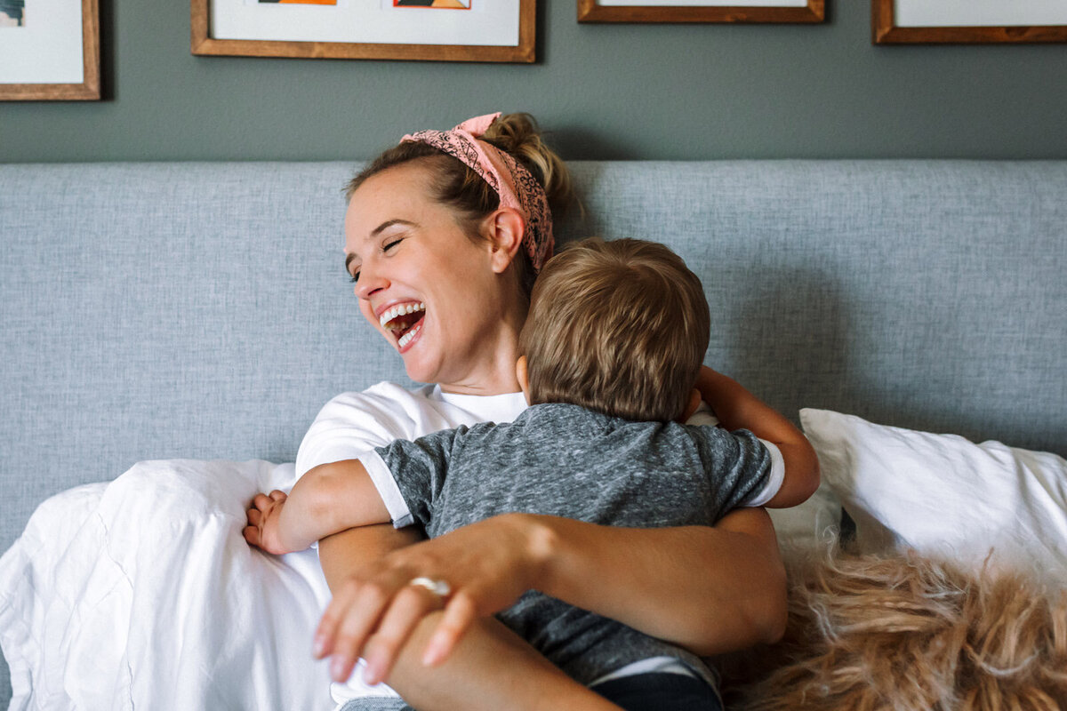 mom leaning up in bed as toddler throws himself on her as she laughs