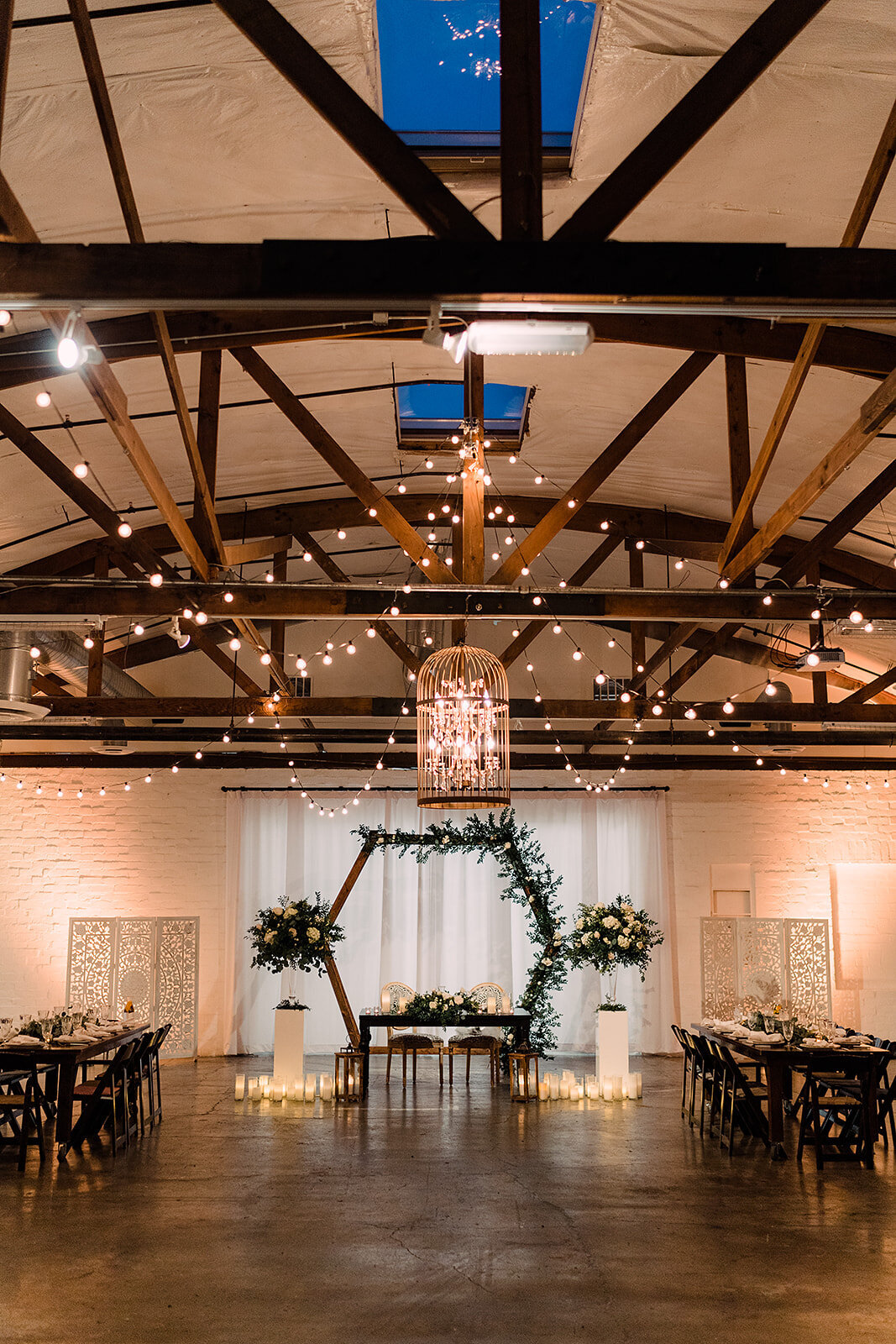 The wedding venue features wooden beams, string lights, and a chandelier hanging above the sweetheart table framed by greenery and candles, creating a romantic and rustic ambiance.
