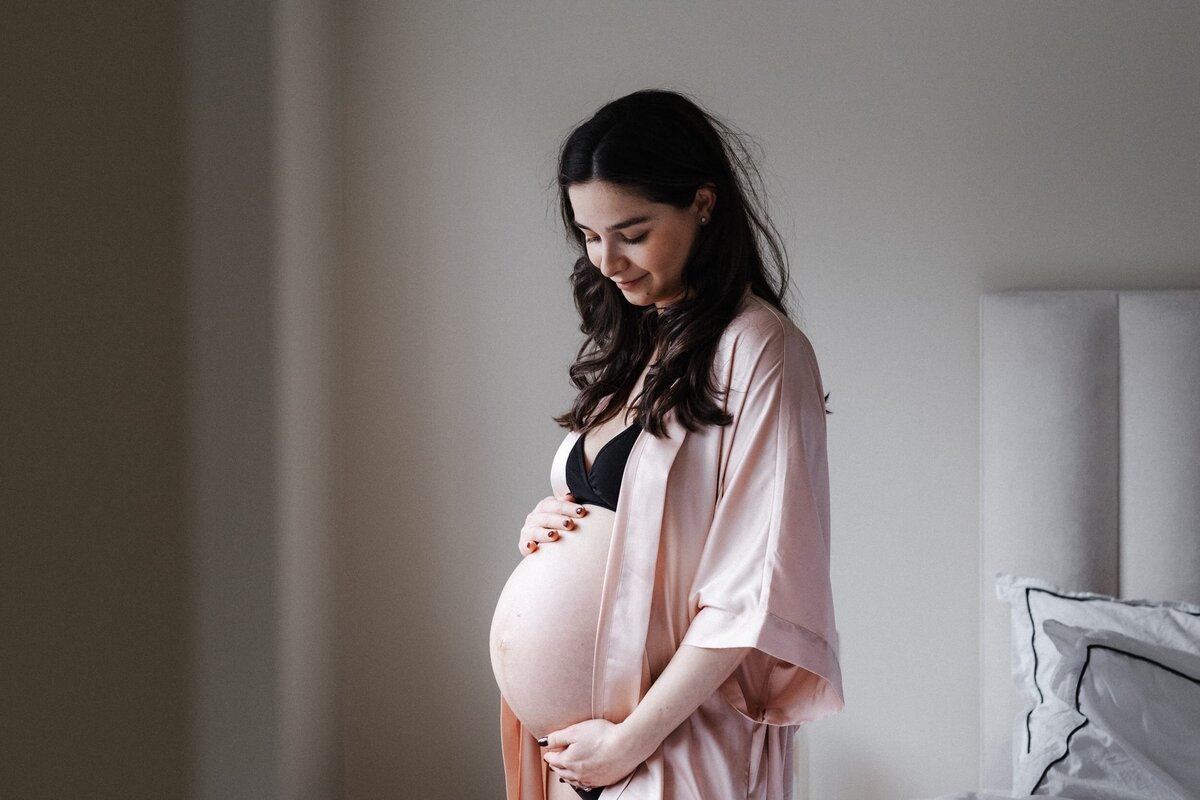Brown haired woman wearing pink silk gown holds her baby bump in her bedroom