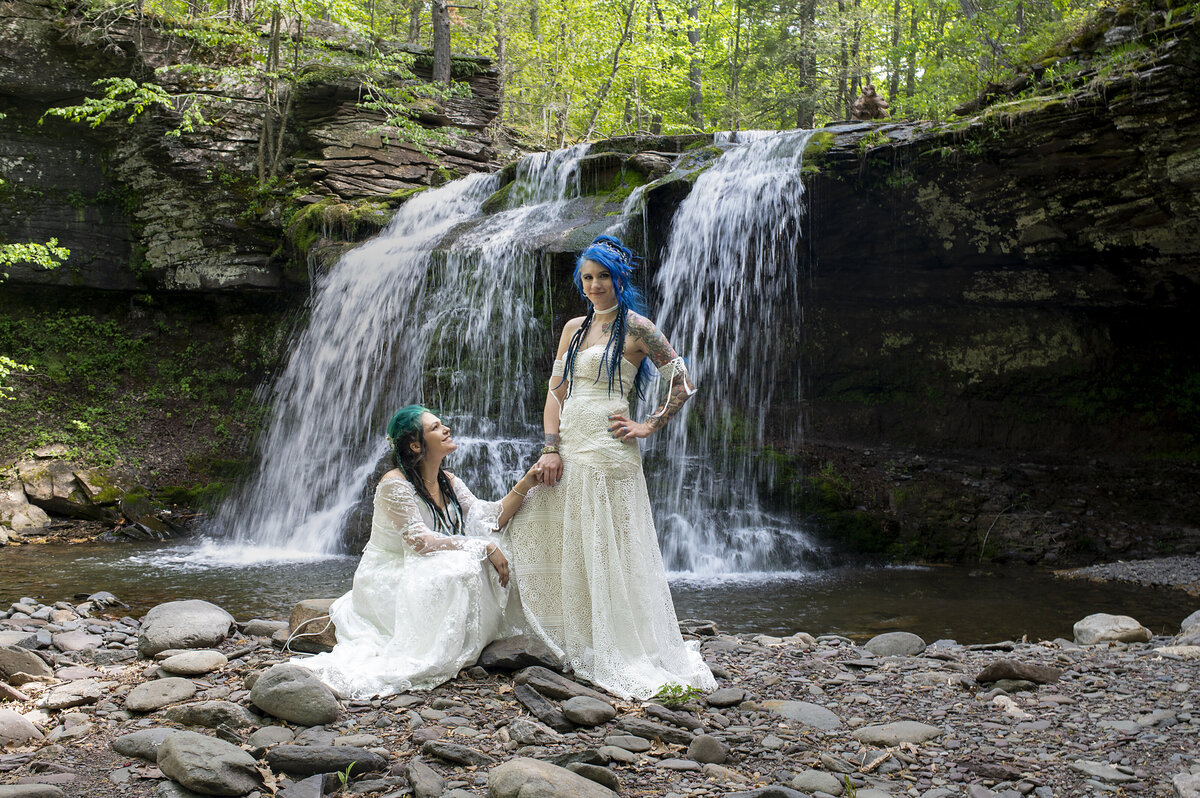 A person squatting down and holding their partner's hand as they're both in wedding dresses 