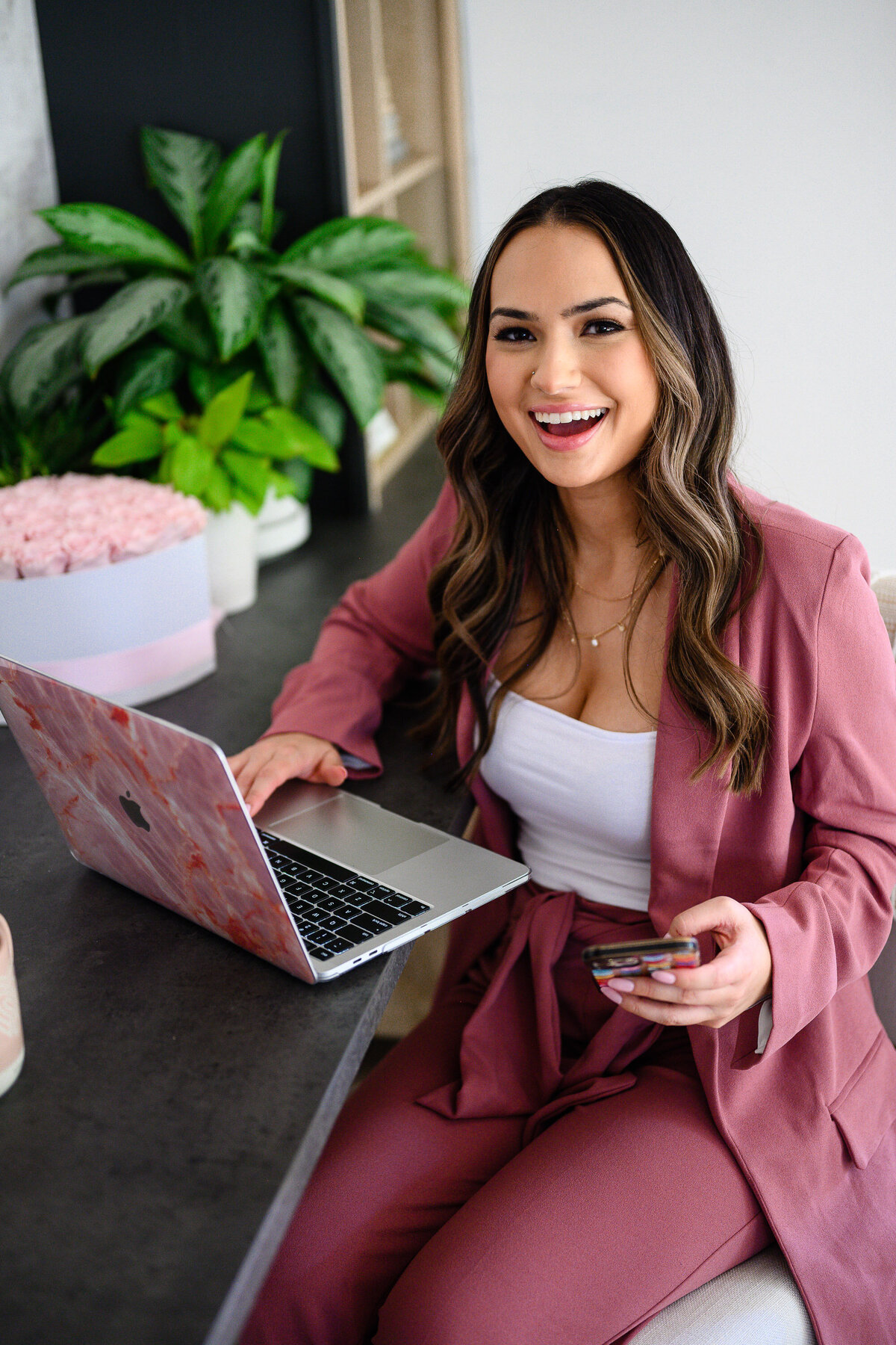 brand photography in studio with business woman in a mauve power suit sitting on a desk and leaughing as she works on her computer captured by denver commercial photographer
