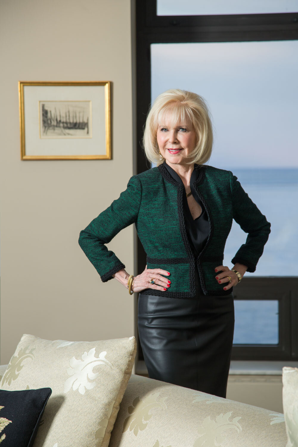 Woman in green blazer and leather skirt standing by a window with a lake view